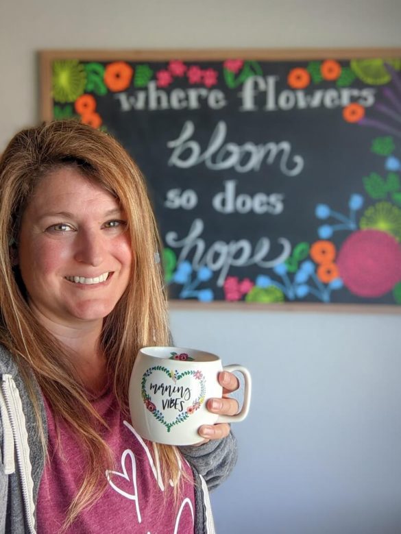 A mom holding a cup of coffee with a chalkboard in the background that reads "where flowers bloom so does hope".