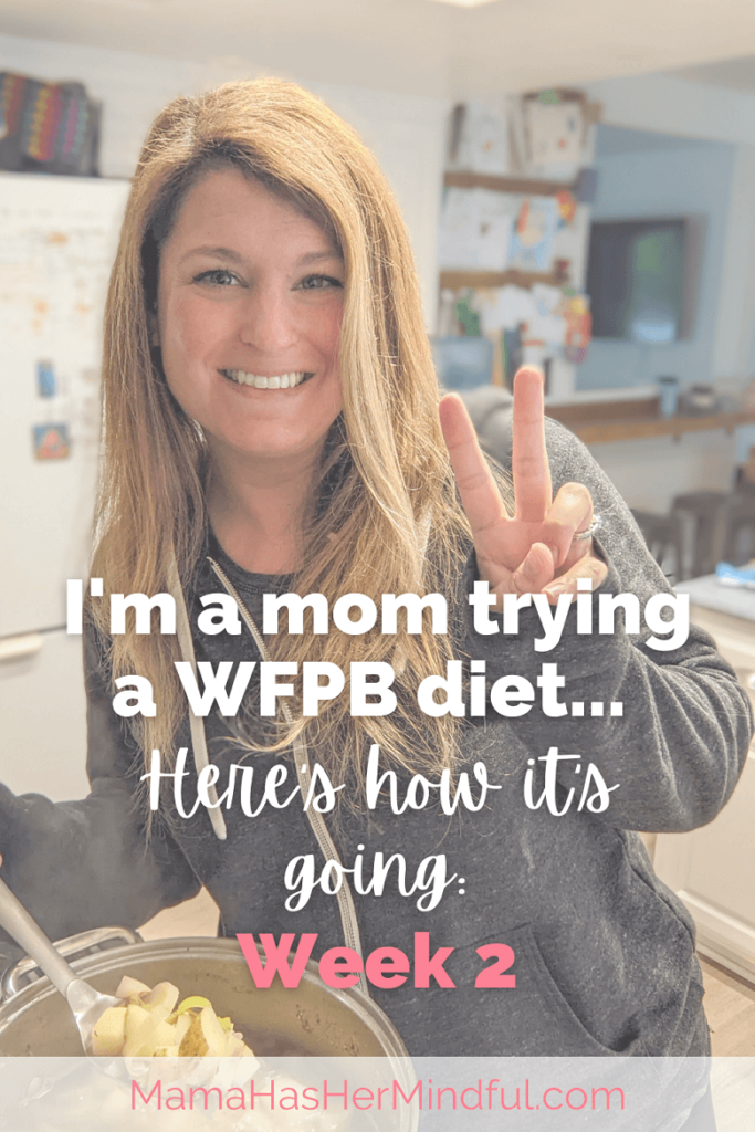 A woman with one hand holding a spoon and stirring a pot of soup and vegetables on the stove and holding up "two" or a peace sign with her other hand. Her kitchen is in the background. The text over the image reads "I'm a mom trying a WFPB diet... Here's how it's going: Week 2" and the URL Mama Has Her Mindful dot com is at the bottom of the image.