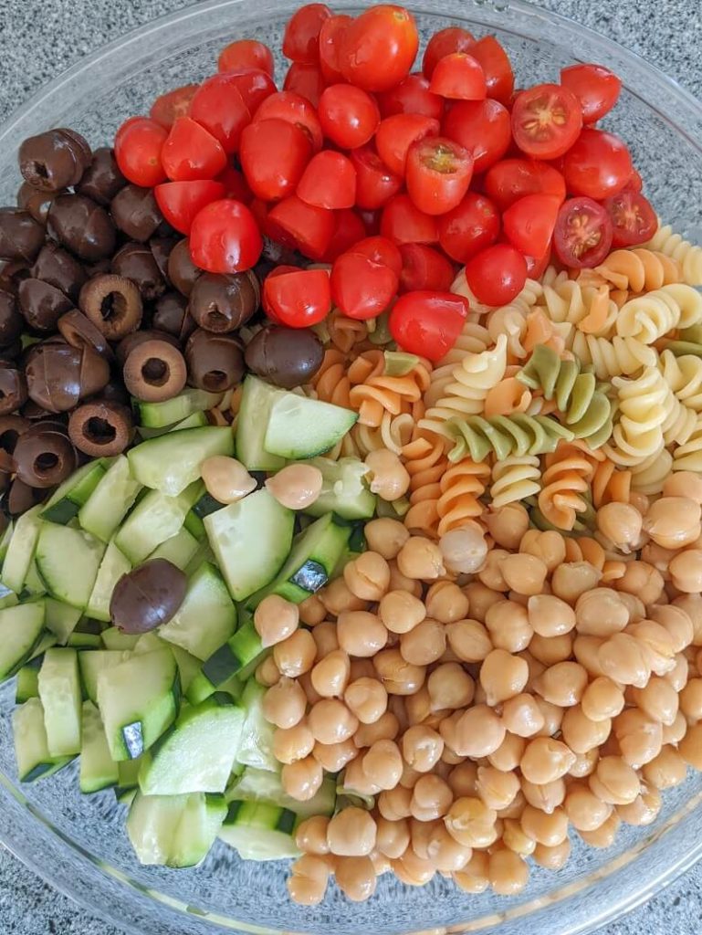 Ingredients for vegan pesto pasta salad that include pasta, garbonzo beans, cucumbers, black olives, and grape tomatoes in a bowl.